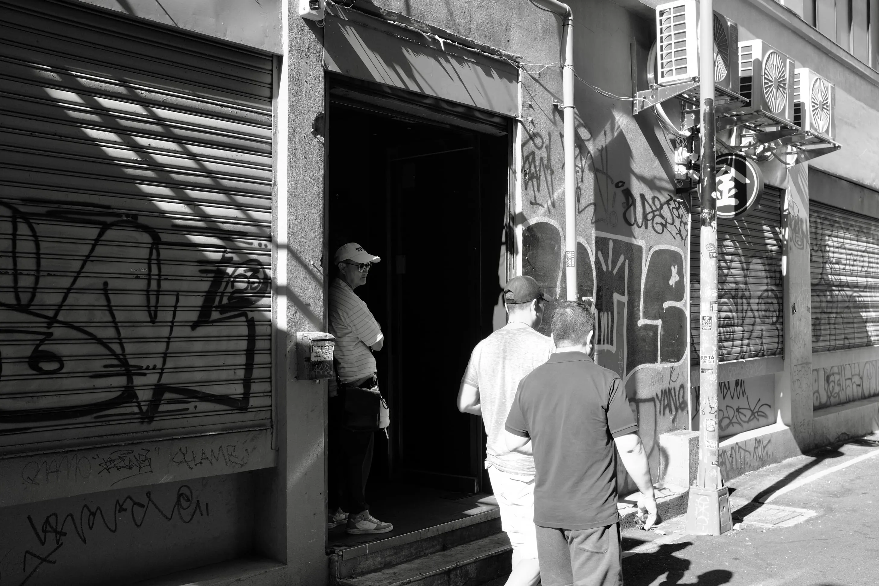 塗鴉鐵門與深色門口旁男子站在陰影裡，兩名行人從前方經過，365 Project September 24