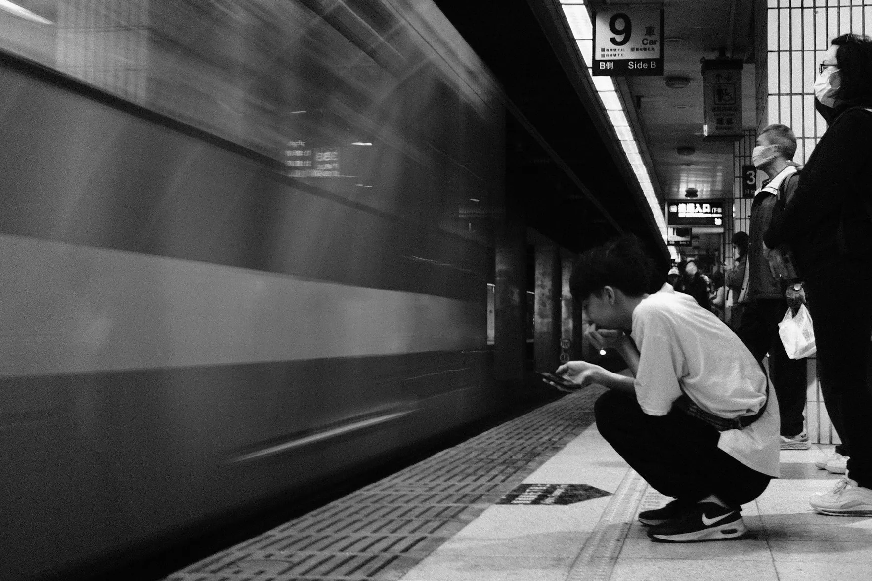列車以動態模糊駛入月台，前景男子蹲在安全線後低頭看手機，後方旅客站立等候，365 Project September 27