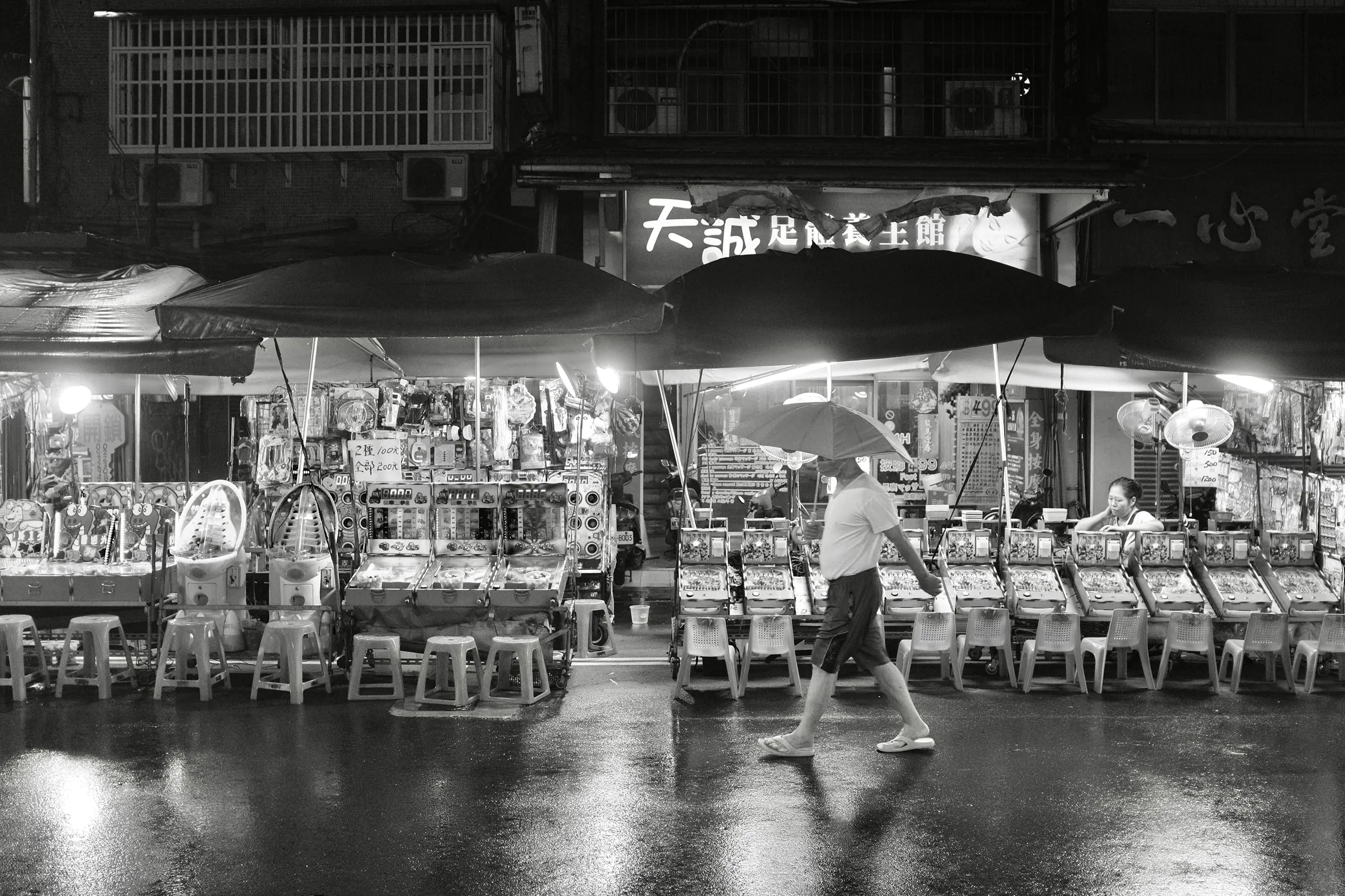 雨夜街邊攤位前一名行人走過，明亮棚燈下排列著椅子與商品，365 Project October 12