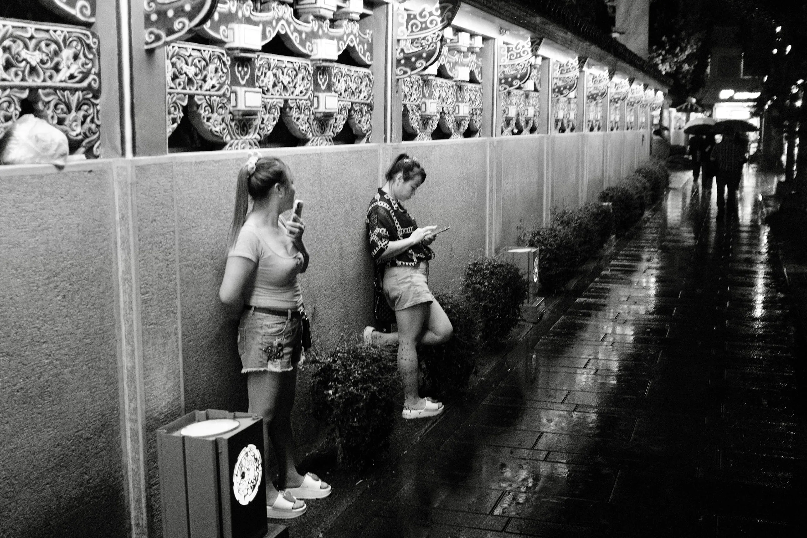 雨後人行道旁兩人靠在裝飾牆前，各自低頭看手機，365 Project October 14
