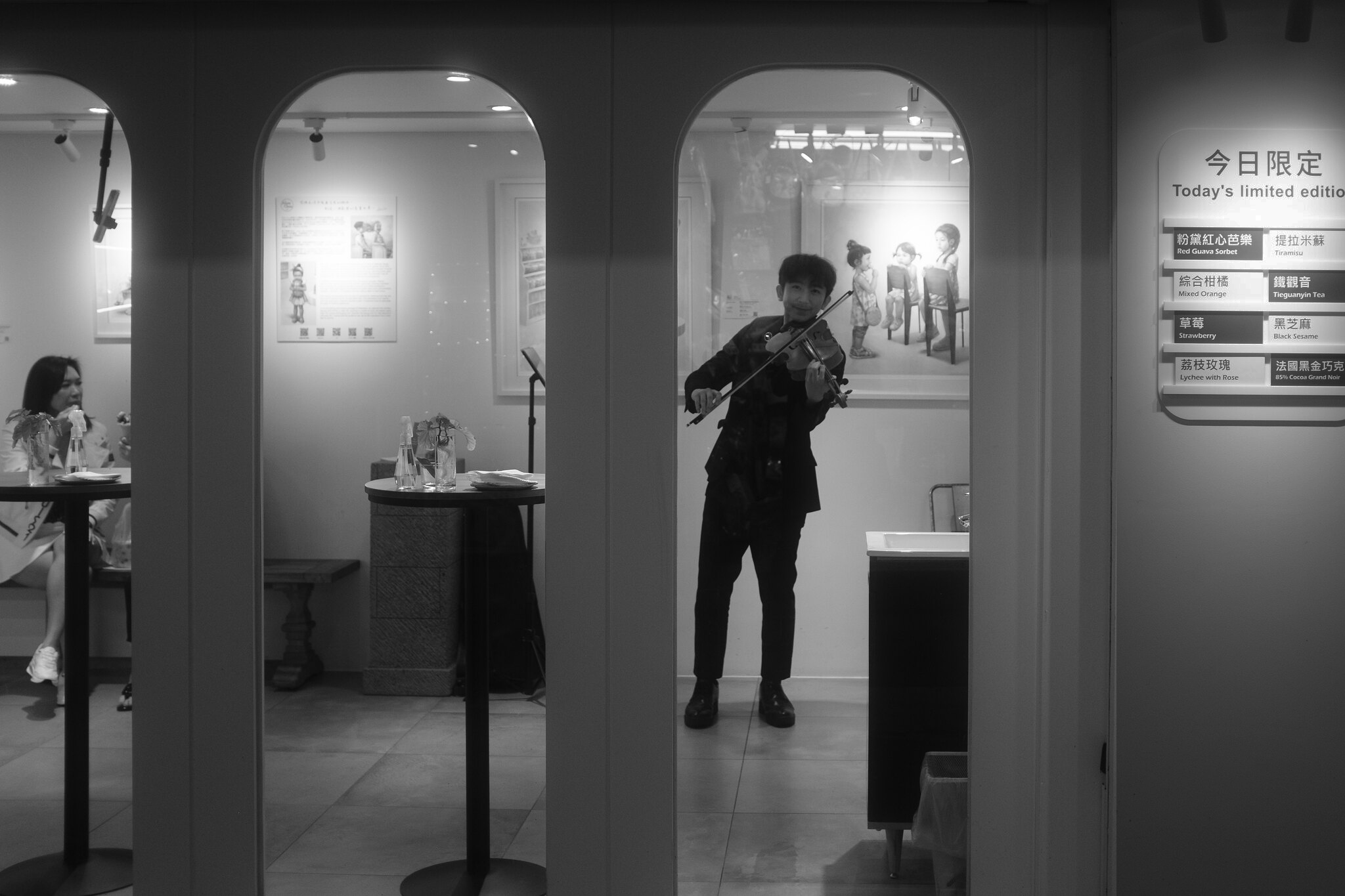 Black and white East District storefront scene with a violinist performing behind arched windows beside a limited edition dessert menu