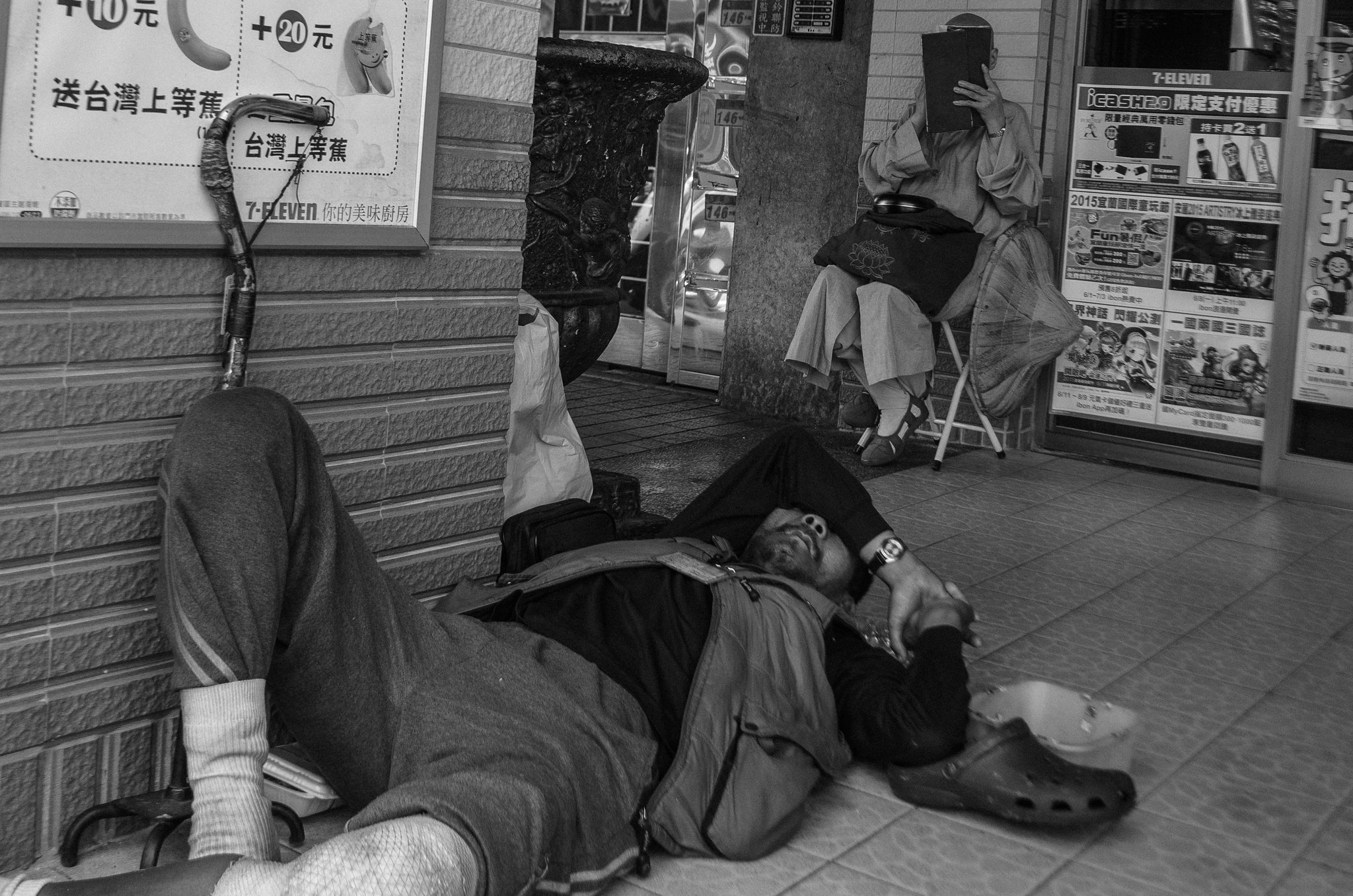 A monk sits on a folding chair reading a book under a shop sign, while a person rests on the ground nearby in a gritty urban street scene