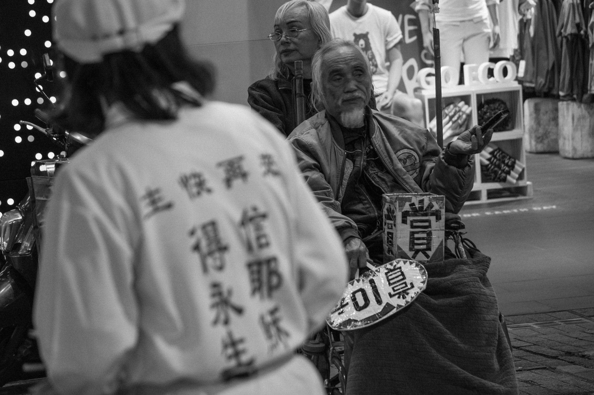 A street preacher with text on their shirt reading 'The Lord is coming soon, believe in Jesus for eternal life' stands opposite an elderly man sitting in a wheelchair