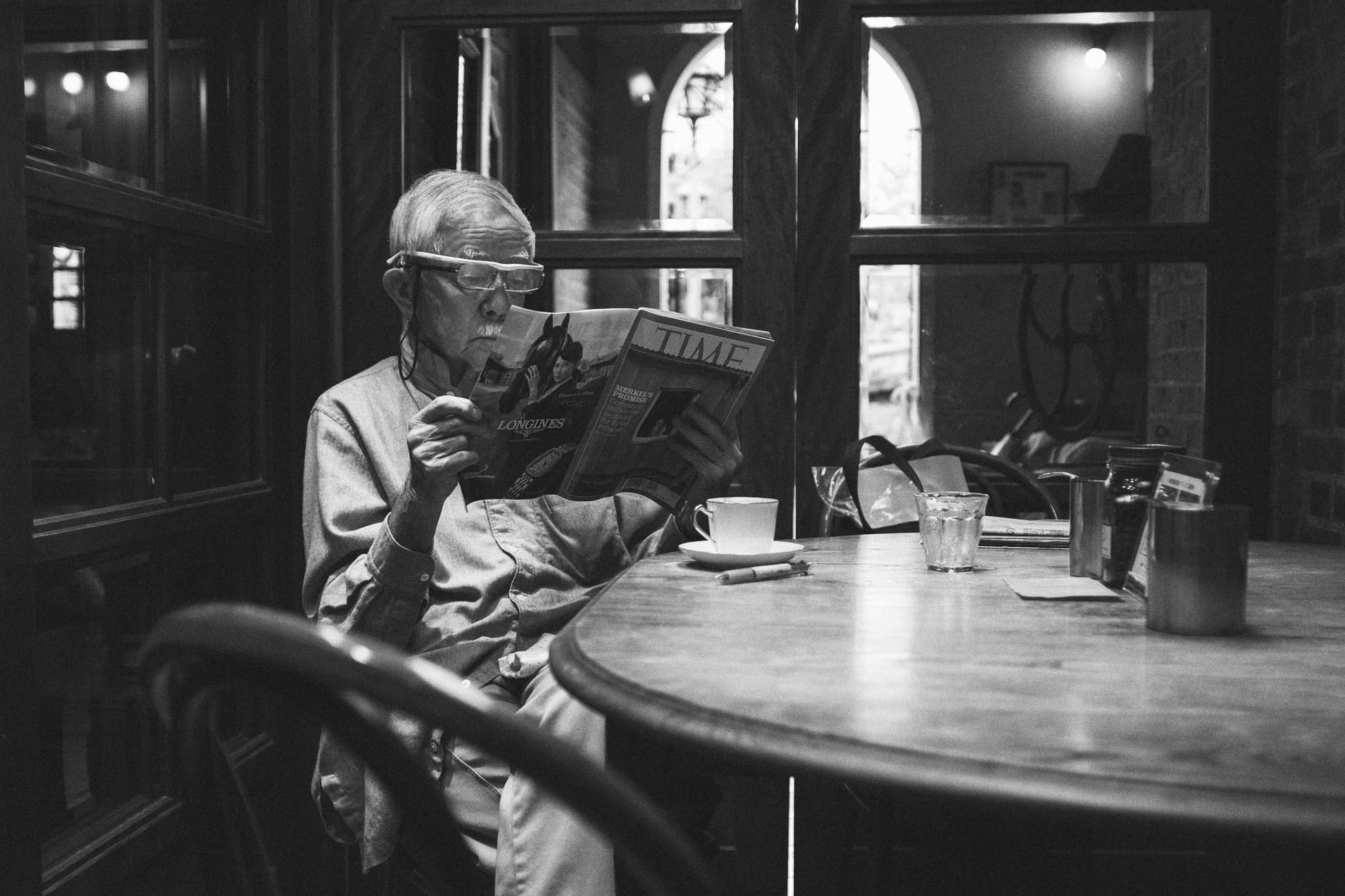 一名年長男子在咖啡館木桌旁閱讀雜誌，窗光落在他的身上