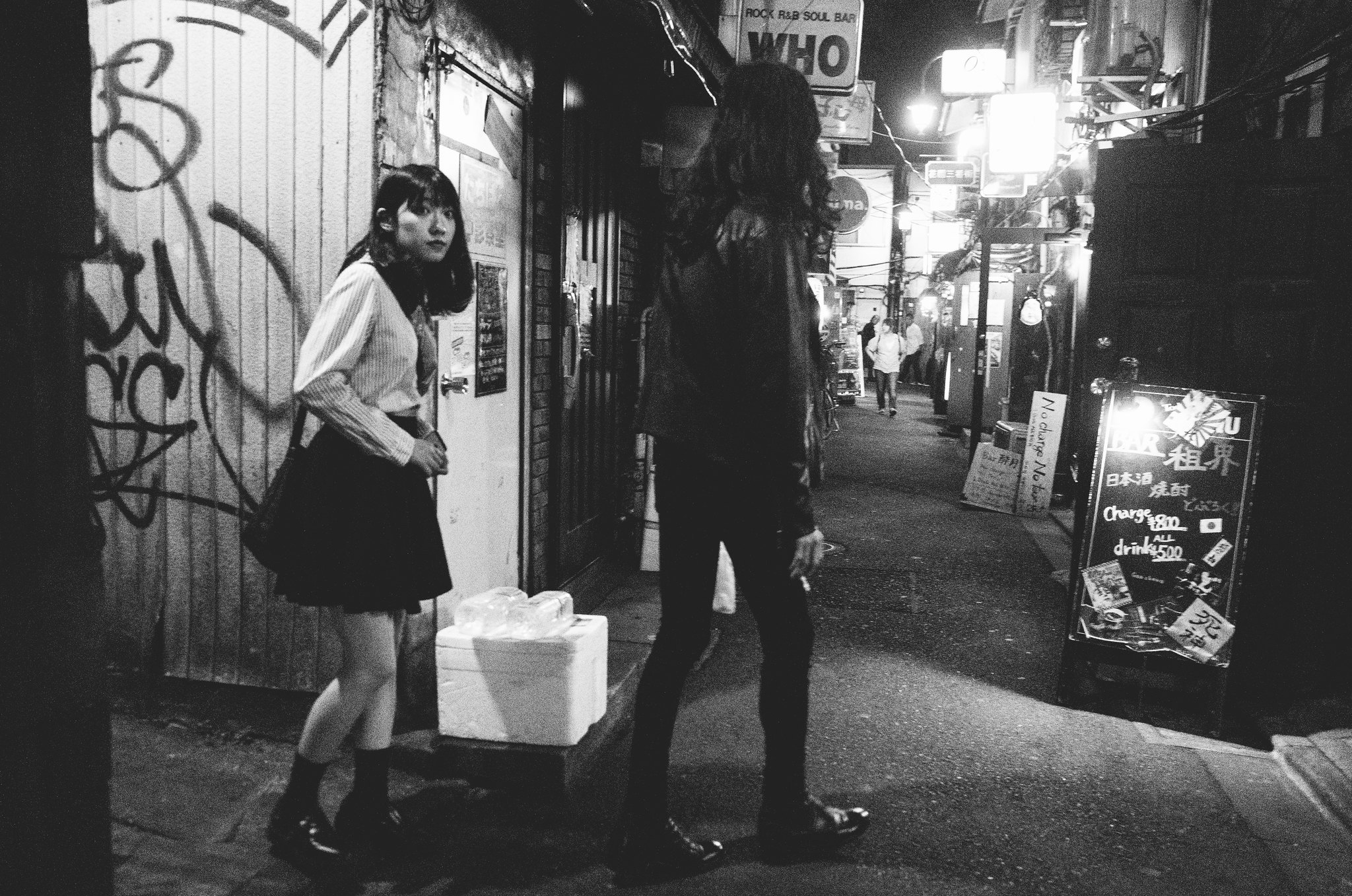 Black and white Japanese alley at night with pedestrians walking past lit storefronts
