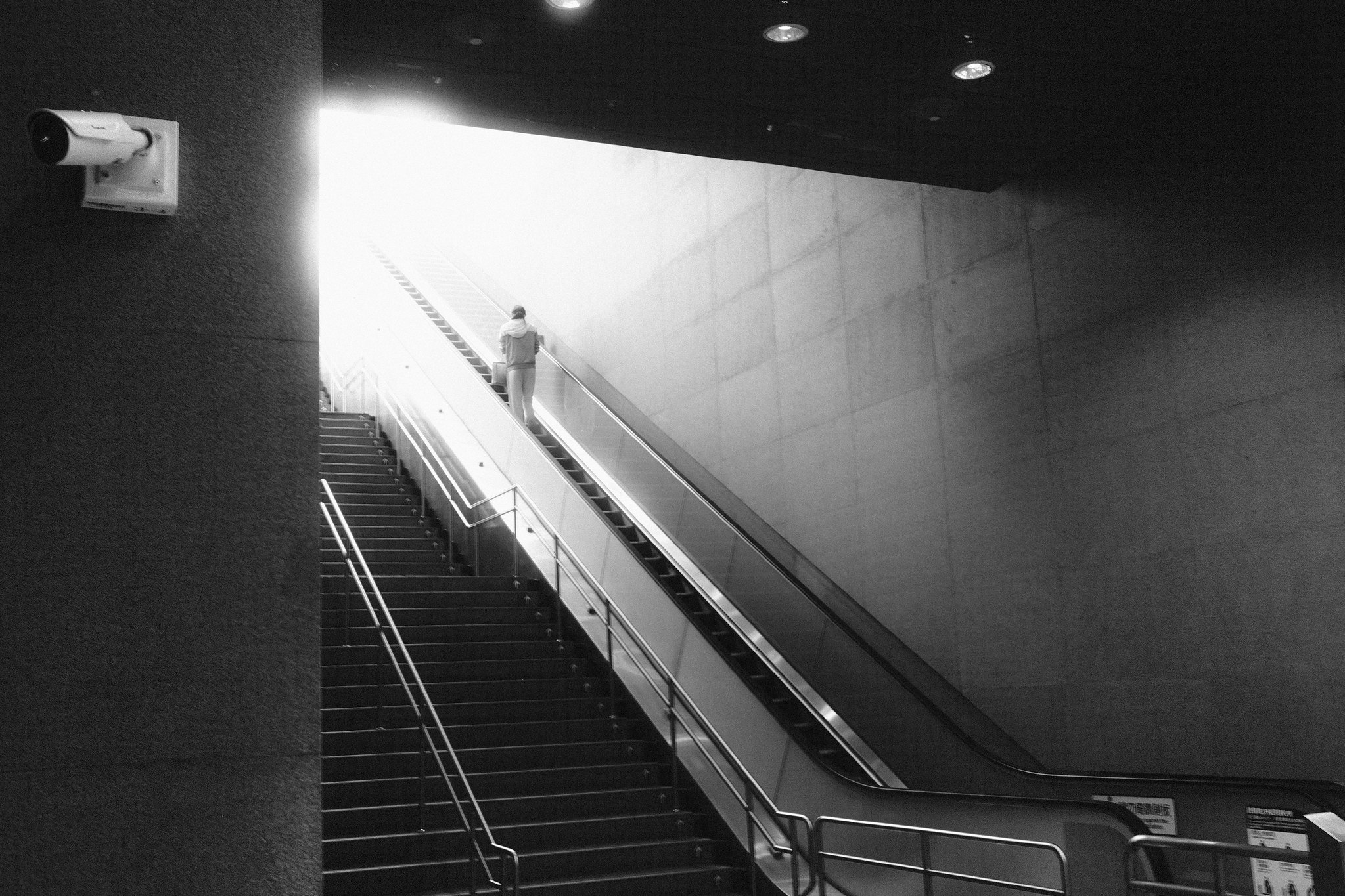 Black and white urban scene of a lone figure riding an escalator toward harsh threshold light in Taiwan