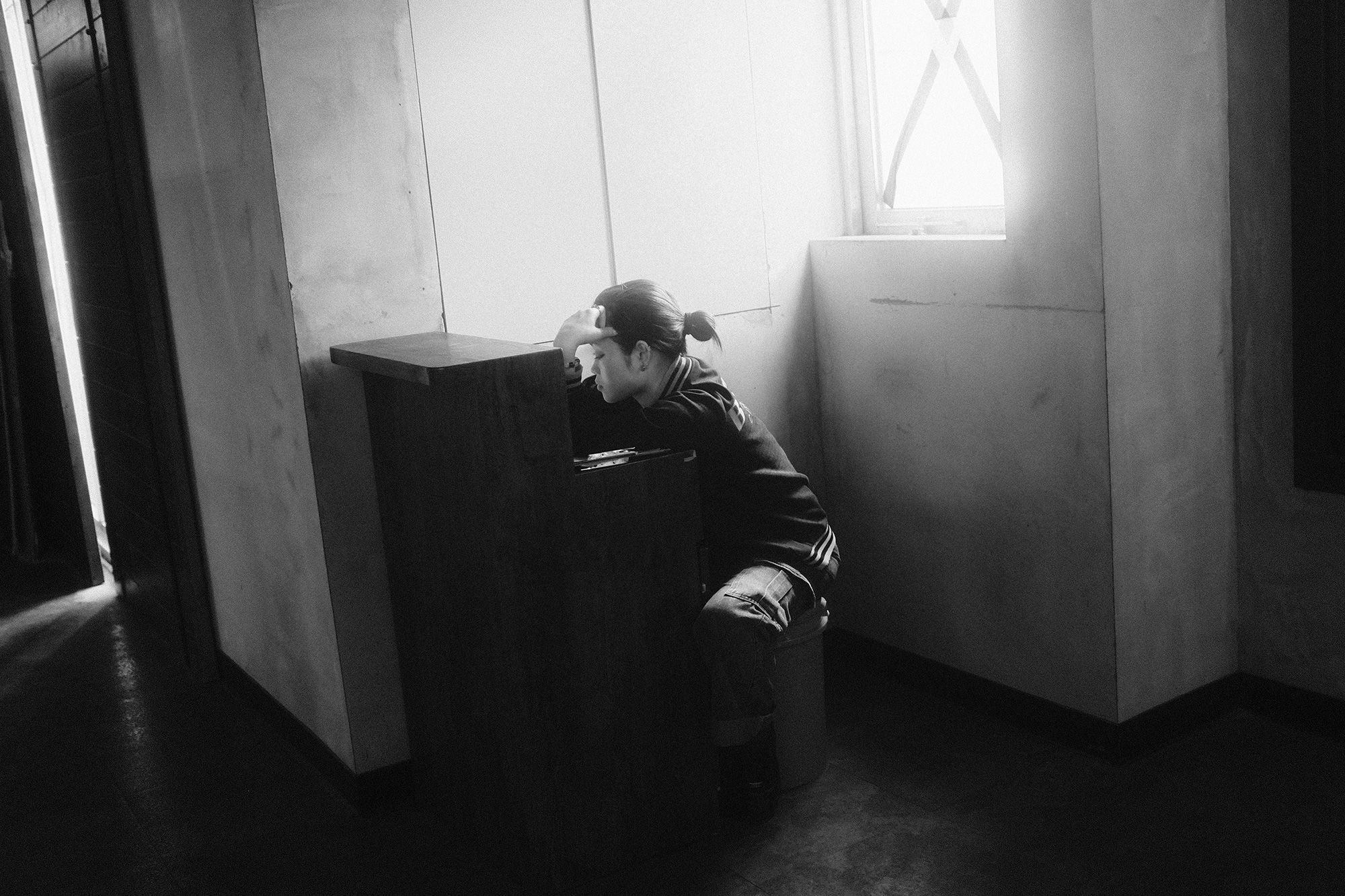 Candid black and white street photo of a worker resting at a small counter, framed by urban fatigue and quiet texture in Taiwan