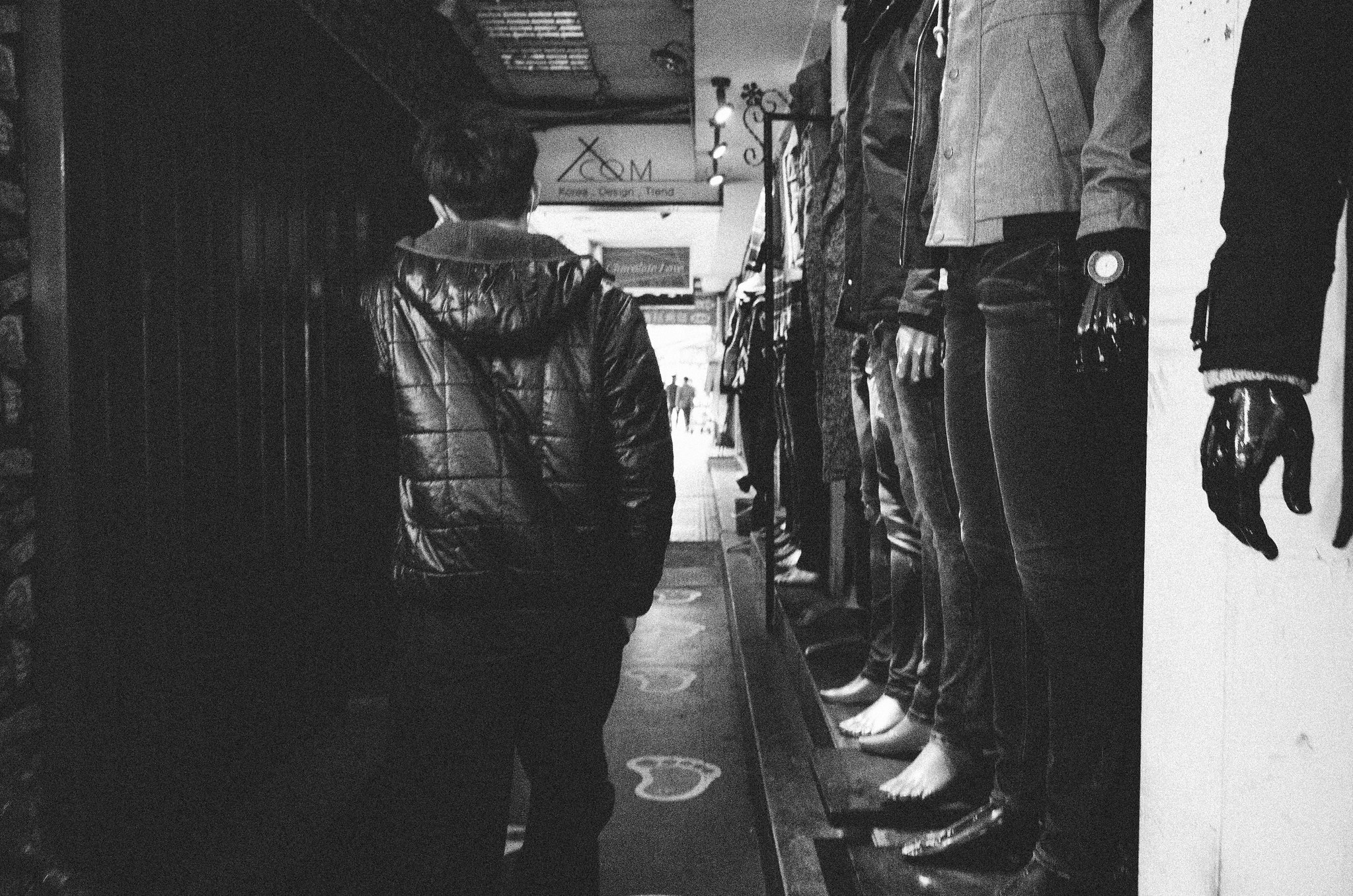 Black and white street scene of a person in a hooded jacket walking past shop window mannequins