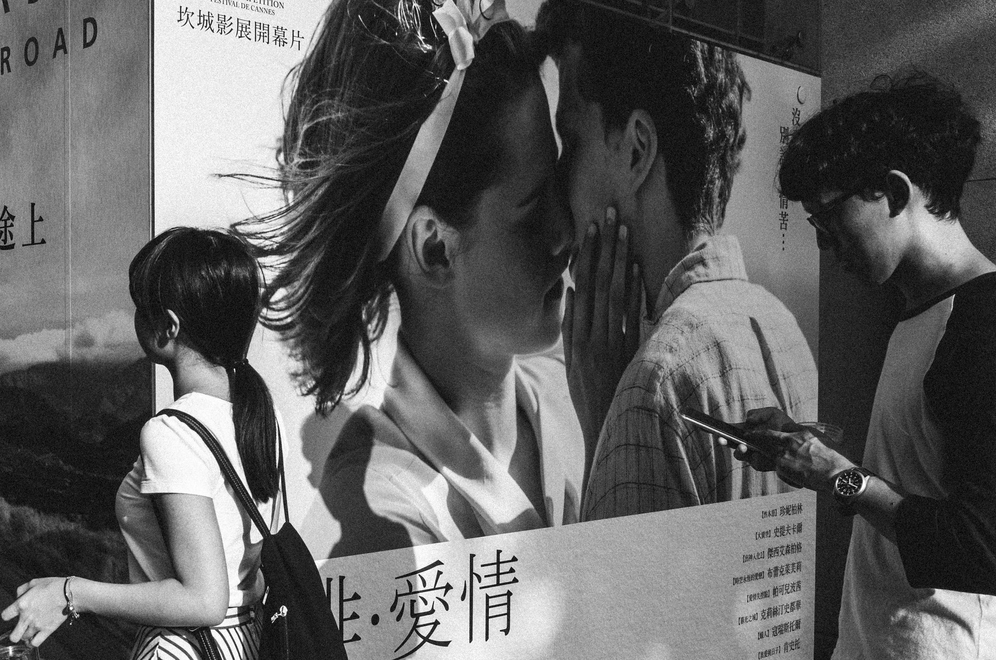 Black and white street photo showing the juxtaposition of a man standing in front of a romantic movie poster