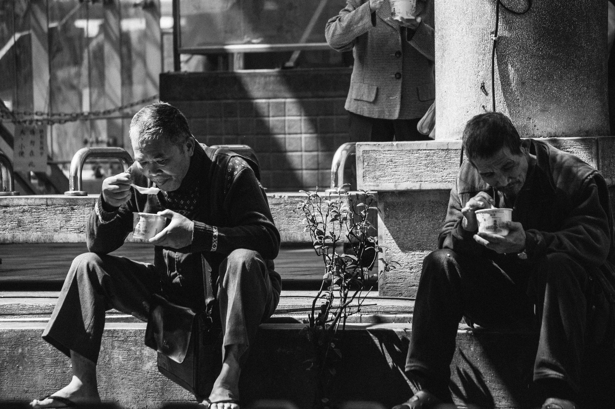Black and white street photograph of two men eating a street-side meal on the steps in Wanhua, Taipei