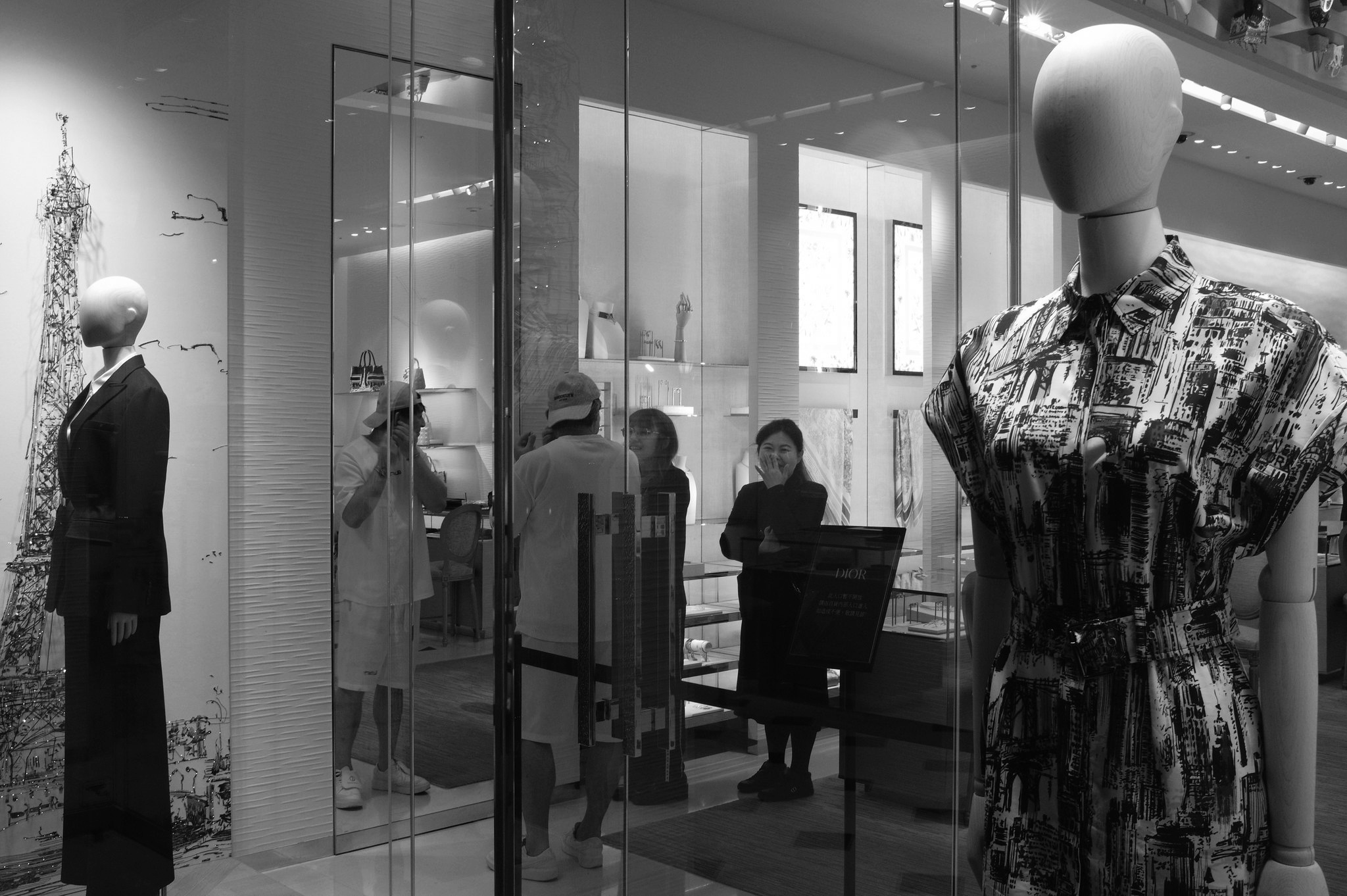 Black and white Xinyi luxury storefront scene with mannequins, glass reflections, and people gathered inside a boutique