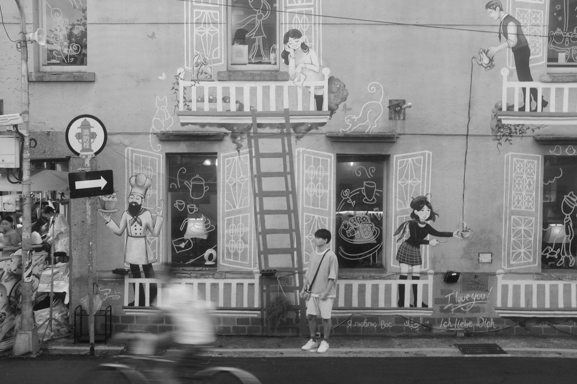 Black and white Zhongshan, Taipei street scene with a painted cafe mural, a standing pedestrian, and a blurred scooter crossing in front