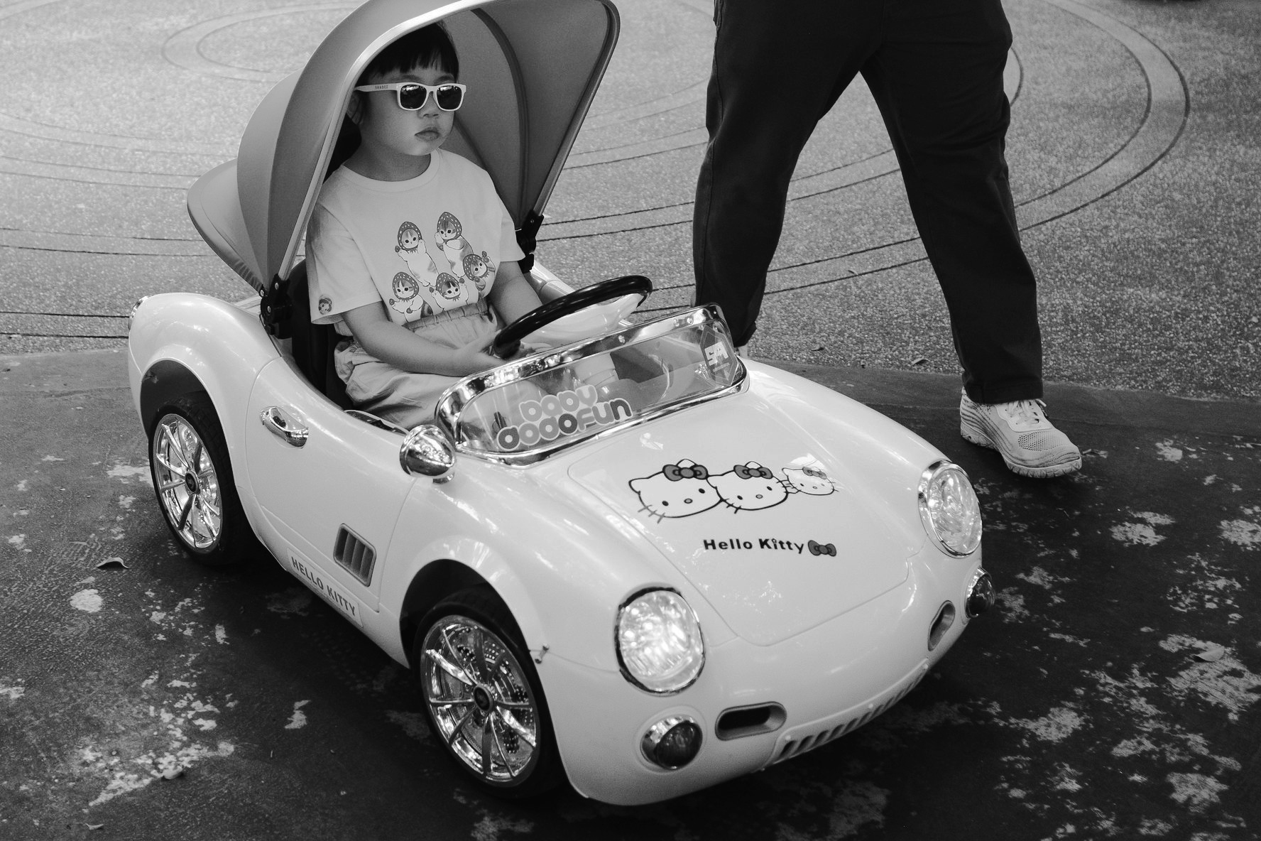 Black and white Zhongshan, Taipei street photograph of a child wearing sunglasses seated in a Hello Kitty ride-on car while an adult walks beside it